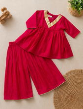 Load image into Gallery viewer, A two-piece baby outfit set in bright pink featuring a long-sleeve peplum-style top with a gold-trimmed V-neck and matching wide-leg pants, laid flat on a beige background with styled props.